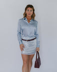 Woman wearing a light blue satin shirt and silver sequin skirt, holding a brown handbag against a white background.
