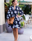 Woman in a floral dress standing outdoors with plants and chairs in the background