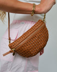Brown woven handbag held by a person wearing a white top and pink pants.