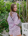 Woman wearing a purple patterned dress standing outdoors with greenery in the background, back view