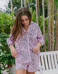 Woman wearing a purple paisley outfit standing outdoors with greenery in the background front view