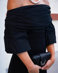Person wearing a black off-shoulder dress holding a black clutch against a neutral background, close up