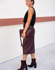 Woman in black top and brown leather skirt with matching boots against a white wall, side/back view