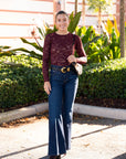 Woman in maroon top and blue jeans standing outdoors with plants and a building in the background