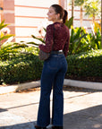 Woman in a red top and blue jeans walking outdoors with plants and a building in the background, back