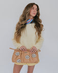 Woman wearing yellow sweater, holding a beige clutch with floral embellishments against a plain background