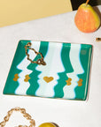 Rectangular clutch with green and white wave pattern and gold chain on a white surface with a pear and gold bracelet.