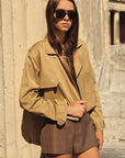 Woman wearing a beige jacket and sunglasses standing against a stone wall.