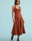 A woman modeling a rust-orange two-piece knit set consisting of a sleeveless scoop-neck top and a matching A-line midi skirt, paired with dark socks and brown lace-up shoes, against a solid light blue studio background.