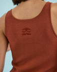Close-up back view of a person wearing a rust-colored ribbed tank top with a tonal embroidered logo on the upper back, against a light blue background.
