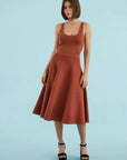 A woman modeling a sleeveless rust-brown midi dress with a scoop neckline and flared skirt, paired with black platform sandals, against a solid light blue studio background.