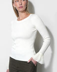 A woman modeling a white long-sleeve ribbed knit top with textured lace flared cuffs, paired with dark olive-brown shorts, against a plain light gray wall.