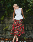 Woman wearing a white top and red floral skirt standing against a stone wall with greenery.