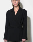 A woman modeling a black wrap-style blazer with a self-tie waist belt, paired with gray textured trousers, against a plain off-white background.