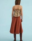 A woman shown from the back wearing a sleeveless spaghetti-strap top with a multicolor floral print and a rust-colored A-line midi skirt, paired with brown knee-high boots, against a solid light blue background.