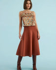 A woman modeling a sleeveless top with a multicolor floral pattern and a rust-colored satin midi skirt, paired with brown knee-high boots, against a solid light blue background.