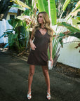 A woman stands outdoors on a paved path surrounded by large tropical green leaves and a white fence, wearing a dark brown sleeveless mini dress with silver stud detailing, holding a small metallic clutch, and wearing white heeled sandals.