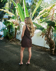 A woman with long wavy blonde hair stands outdoors on a paved path wearing a sleeveless dark brown mini dress, photographed from behind. Large green banana plant leaves and a white fence are visible in the background.