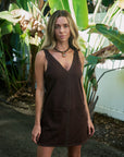 A woman wearing a dark brown sleeveless mini dress with silver stud detailing, standing outdoors in front of tropical green plants.