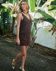 A woman wearing a sleeveless brown mini dress with decorative stitching, walking outdoors on a paved path surrounded by tropical banana leaves and a white fence.