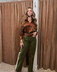 A woman modeling a two-piece outfit consisting of a brown satin long-sleeve blouse and olive green wide-leg trousers with a white side stripe, standing in front of brown velvet curtains.