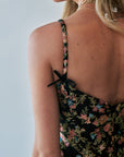 Close-up back view of a woman wearing a floral spaghetti-strap dress with black ribbon ties at the shoulders, against a light gray background.