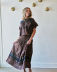 A woman modeling a matching brown two-piece set consisting of a short-sleeve ruffled top and a pleated maxi skirt with dark blue horizontal stripes, standing in a room with a white wall and light wood floor.