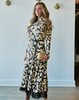 A woman stands indoors wearing a long-sleeve midi dress with a black base and a cream and yellow floral print, along with a matching black belt and fringe hem. She is holding a small beige clutch and wearing white heeled sandals.
