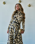 A woman modeling a long-sleeve high-neck maxi dress with a black and beige floral pattern, holding a small gold clutch against a light blue wall with gold wall decor.