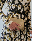 A close-up of a person wearing a long-sleeve dress with a black, olive green, and cream abstract pattern, holding a woven straw clutch bag with a gold chain strap.