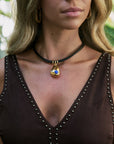 A close-up of a woman wearing a black choker necklace with a gold teardrop pendant featuring a blue evil eye design, paired with a brown studded top.