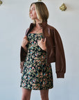 A woman wearing a brown faux-leather bomber jacket over a black floral strapless dress, standing against a white wall with gold wall sconces.