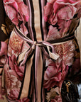 A close-up of a floral dress or garment featuring large pink and mauve peony-like flowers over a black base with vertical pink and beige stripes, tied at the waist with a matching satin ribbon. A small black handbag is partially visible at the lower right.
