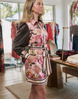 A woman models a sleeveless floral mini dress with a striped tie belt over a dark brown long-sleeve shirt in a boutique setting.