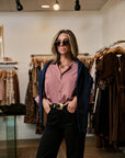 A woman posing in a clothing boutique wearing a pink and white striped button-down shirt, a navy blue zip-up cardigan, black trousers, and dark sunglasses.