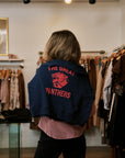 A person viewed from behind wearing a navy blue cropped hoodie with 'THE GREAT PANTHERS' and a panther graphic in red on the back, standing inside a clothing boutique with racks of garments in the background.