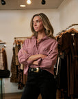 A woman stands in a clothing boutique wearing a pink and white vertically striped button-down shirt with black pants and a gold belt buckle.