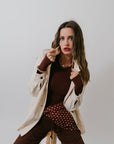 Woman in ivory jacket, brown bodysuit, polka dot scarf and brown pants against a plain background.