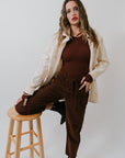 Woman in brown outfit sitting on a wooden stool against a white background