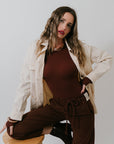Woman in a beige jacket, Ballet Crewneck Bodysuit and brown pants sitting on a stool against a plain background