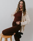 Woman sitting on a wooden stool wearing a brown outfit with a Charlie Chore Coat against a plain background