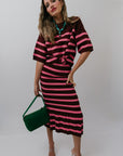 Woman wearing a maroon and pink striped set holding a green handbag against a white background, front