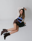 Woman sitting on a white floor wearing a beige blazer, striped top, brown skirt, and brown boots.