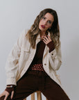 Woman sitting on a stool wearing a Charlie Chore Coat and brown pants against a plain background