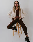 Woman sitting on a stool wearing a beige jacket, brown pants, and black shoes against a white background