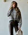 A woman models a gray knit sweater with a Fair Isle pattern in burgundy and cream, paired with dark brown leather pants and holding a beige handbag against a light gray wall.