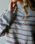 A close-up of a woman wearing a light gray knit sweater with burgundy geometric Fair Isle patterns across the chest and sleeves.