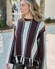 A woman wearing a knit sweater with vertical stripes in burgundy, cream, and black, featuring a fringed hem and long sleeves, standing outdoors.