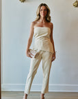 A woman models a cream-colored strapless top and matching cropped trousers, holding a small patterned clutch and wearing nude high-heeled sandals against a white wall with gold wall decor.