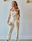 A woman models a matching cream-colored two-piece set consisting of a strapless top and cropped trousers, styled with a matching clutch and nude heels against a light gray wall.
